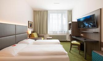 A modern hotel room with two single beds and a desk. The room is bright and friendly, with a large window and a TV on the wall.