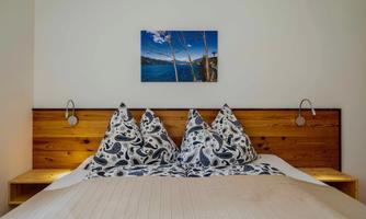 A cozy bedroom with a wooden headboard and decorative pillows. On the wall hangs a painting of a picturesque landscape with water and mountains.