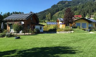 A green meadow with two houses that have solar panels on the roof. In the background, gentle hills and trees can be seen.