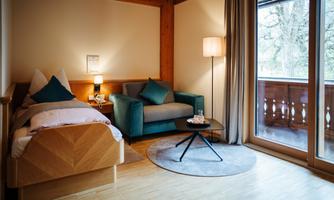 A modern room with a single bed and a seating area. Large windows offer a view of a balcony and plenty of natural light.