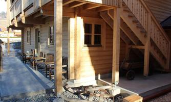 A wooden house with a staircase and a covered area. On the terrace, there are some chairs and tables.