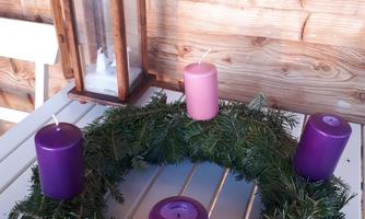 An advent wreath with four candles, surrounded by fir branches. In the background, a wooden lantern is on a table.