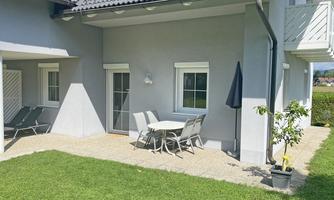 A bright terrace with a table and chairs, surrounded by green lawn. In the background, sun loungers and a parasol can be seen.