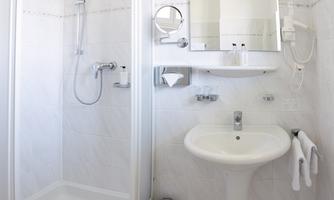 A modern bathroom with a shower, a sink, and a large mirror. The walls are tiled in light colors and there are towels as well as a trash can.