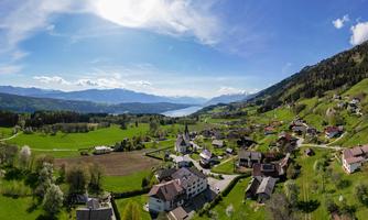 A picturesque landscape with a small village, surrounded by green meadows and mountains. The blue sky and the sparkling lake in the background give the scene a tranquil atmosphere.