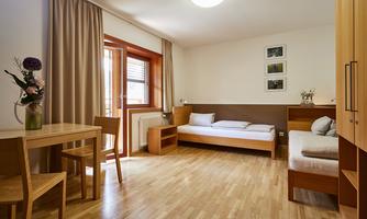 A bright room with wooden floors and modern furniture.  
There is a bed, a table with chairs, and a balcony with a window.