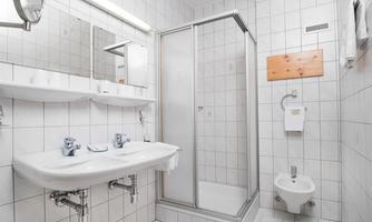 A simple bathroom with white tiles, two sinks, and a shower. There is also a bidet and large mirrors on the wall.
