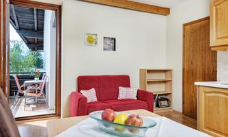 A cozy living room with a red sofa and a small seating area. Through the door, you can see a balcony with a view of the greenery.