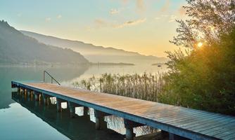 A tranquil lakescape with a wooden dock and gentle hills in the background. The morning sun gently reflects on the water.