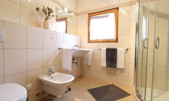 A modern bathroom with cream-colored tiles and a shower. There are towels, a bidet, and a window that lets in natural light.