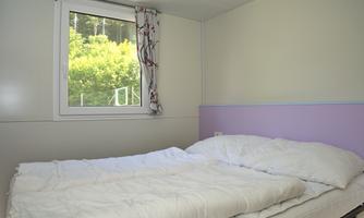 A simple bedroom with a double bed and white blankets. A window allows a view of green trees outside.