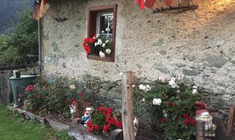 A charming house with a stone wall and colorful flowers in the garden. Pots with red flowers hang in front of the window, and small figures are decorated in the flower bed.
