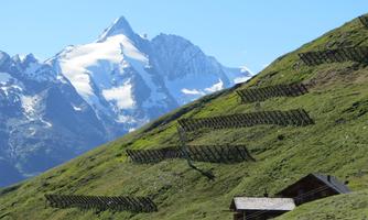 A green mountain landscape with snow-covered peaks in the background. In the foreground, there is a small wooden house.