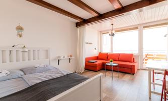 A modern, bright room with a white bed and a red sofa. Large windows provide plenty of daylight and a beautiful view.