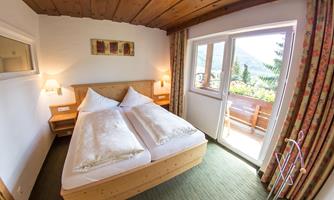 A cozy bedroom with two single beds and wooden furniture. The room has large windows with views of the mountains and a balcony.