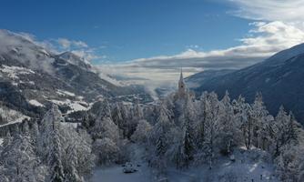 A snowy mountain landscape with tall fir trees and a clear blue sky. In the foreground stands a small church, surrounded by wintry nature.