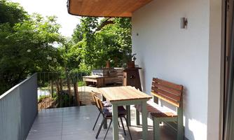 A bright balcony with a wooden chair and a table. The surroundings are green and peaceful, ideal for relaxing.