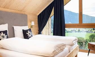 A cozy bedroom with two beds and natural wooden elements. The large window view shows a green landscape and mountains.