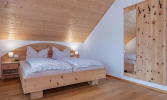 A cozy bedroom with a wooden bed and floral bedspreads. On the wall, there is a mirror and a wooden chest of drawers.