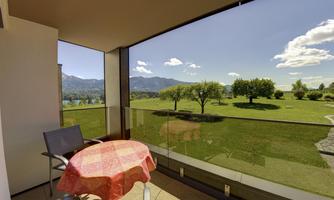A balcony with a round table and a chair. In the background, green meadows and mountains under a blue sky can be seen.
