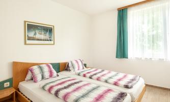 A simple room with two single beds and colorful bedspreads. The room has a window with curtains and a picture on the wall.