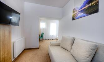 A modern living room with a comfortable, bright sofa and a TV on the wall. In the background, a window and a chair are visible.