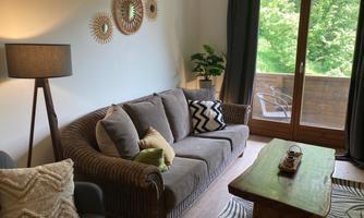 A cozy living room with a beige sofa, a wooden table, and a floor lamp. Large windows offer a view of a green balcony.