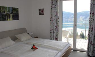 A bright bedroom with two beds and a balcony that offers a view of a lake and the mountains. The decoration is cozy and a few flowers are decoratively placed on the bed.