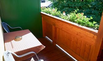 A cozy balcony with a wooden railing and an orange tablecloth. There is a chair and blooming plants in the background.