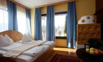 A cozy bedroom with a large window and blue curtains. On the bed are several pillows and a blanket, and a table with fresh fruit stands in the corner.