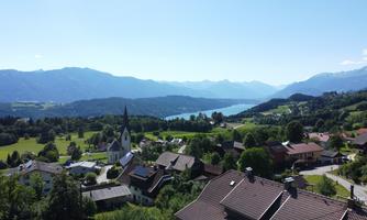 A picturesque landscape with a clear lake and mountains in the background. In the foreground, small houses and a church are visible.
