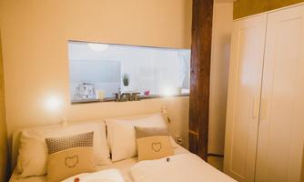 A cozy bedroom with a large bed and white bedding. In the background, a wardrobe and an open kitchen can be seen.