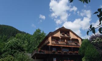 A large wooden house surrounded by green trees and a clear sky. In the foreground, a garden with flowers and plants is visible.