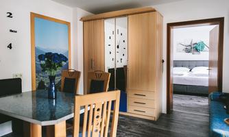 A modern living room with a dining table, wooden chairs, and a wardrobe. In the background, a bedroom with a double bed is visible.