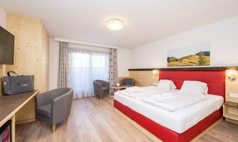 A modern hotel room with a comfortable double bed and wooden paneling. The large window front lets in plenty of daylight, and the room is equipped with a television and a table.