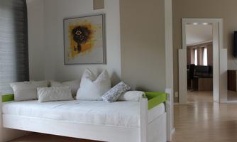 A cozy room with a daybed and several pillows. A colorful artwork hangs on the wall, and another room can be seen in the background.