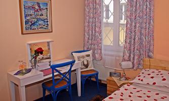 A cozy bedroom with a bed with red dots and colorful curtains. There is a desk with two blue chairs and a painting on the wall.