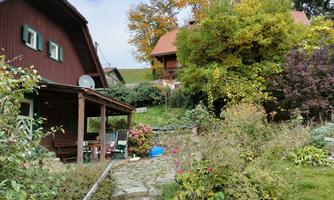 An idyllic garden with colorful flowers and bushes surrounds two wooden houses. The trees display autumn colors and the sky is slightly cloudy.