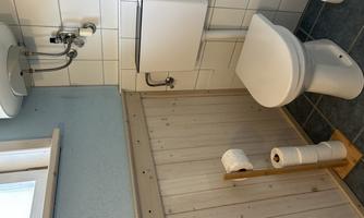 A simple bathroom with a toilet and a wooden shelf for toilet paper. The walls are partially tiled and the room has a bright, friendly ambiance.