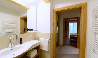A modern bathroom with a sink, mirror, and towels. In the background, a bedroom can be seen.