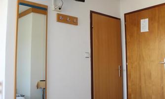 A simple bedroom with two beds and a ceiling lamp. On the wall hangs a television and a mirror next to a wooden board with hooks.