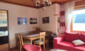 A cozy living room with a red sofa and a wooden dining area. Bright windows provide plenty of daylight and a welcoming atmosphere.