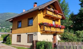 A charming wooden house with balconies decorated with colorful flowers. In the background, mountains and a clear blue sky can be seen.