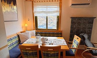 A cozy dining area with a wooden table and chairs. The room has a warm atmosphere with wooden paneling and a window looking outside.