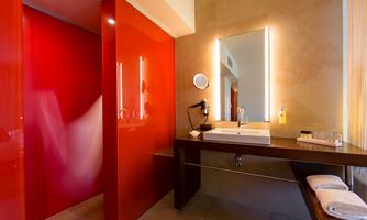 A modern bathroom with red walls and elegant lighting. The vanity is stylishly equipped with towels and toiletries.