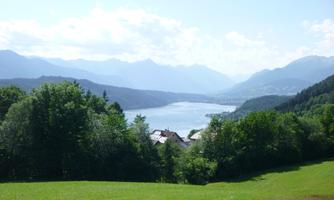 An idyllic landscape with a lake and mountains in the background. The sky is clear and the vegetation is green and lush.