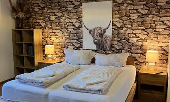 A cozy bedroom with a large double bed and wooden shelves. The walls are decorated with a stone look and a picture of a Highland cow.