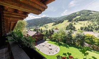 A picturesque mountain landscape with green meadows and trees. In the foreground, flowers can be seen on a balcony and a cozy garden.