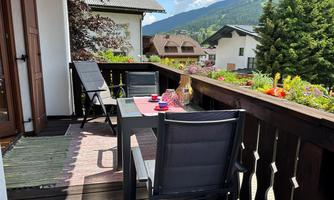A cozy balcony with a table and chairs. In the background, green mountains and colorful flowers can be seen.