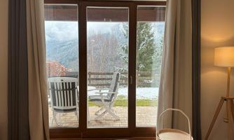 A cozy room with large windows and a view of a terrace. The view shows snow-covered mountains and trees.